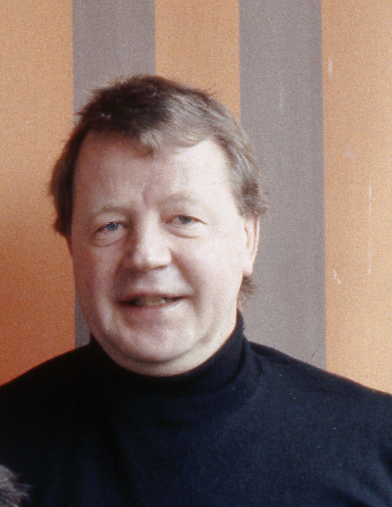 Siegmar Gassert, Porträt aus den 1990er-Jahren: lächelnd, im schwarzen
Rollkragenpullover, vor einer orange-grau gestreiften Wand.